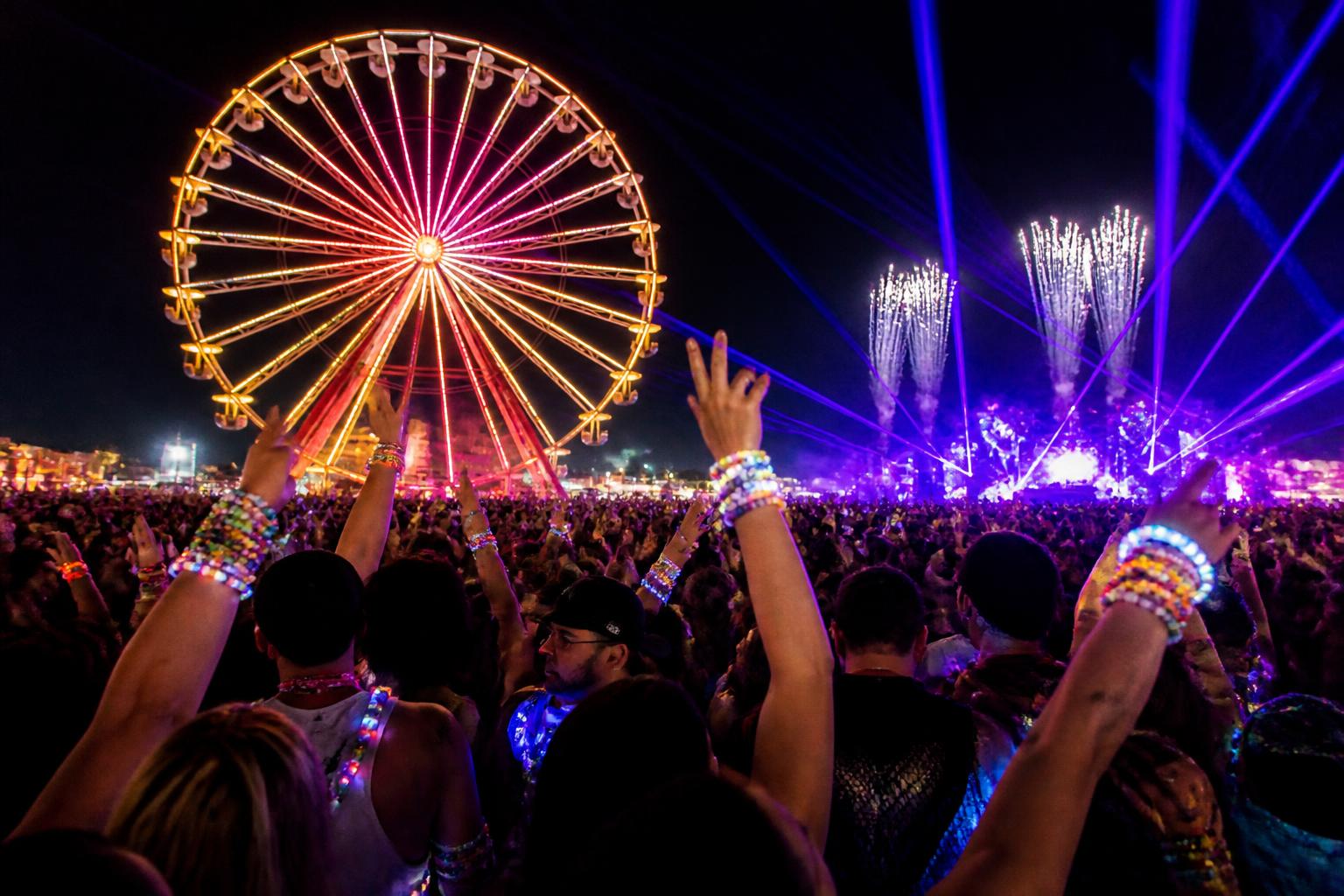 Multitud iluminada con pulseras LED bajo la rueda de la fortuna gigante de EDC, con escenarios y fuegos artificiales de fondo