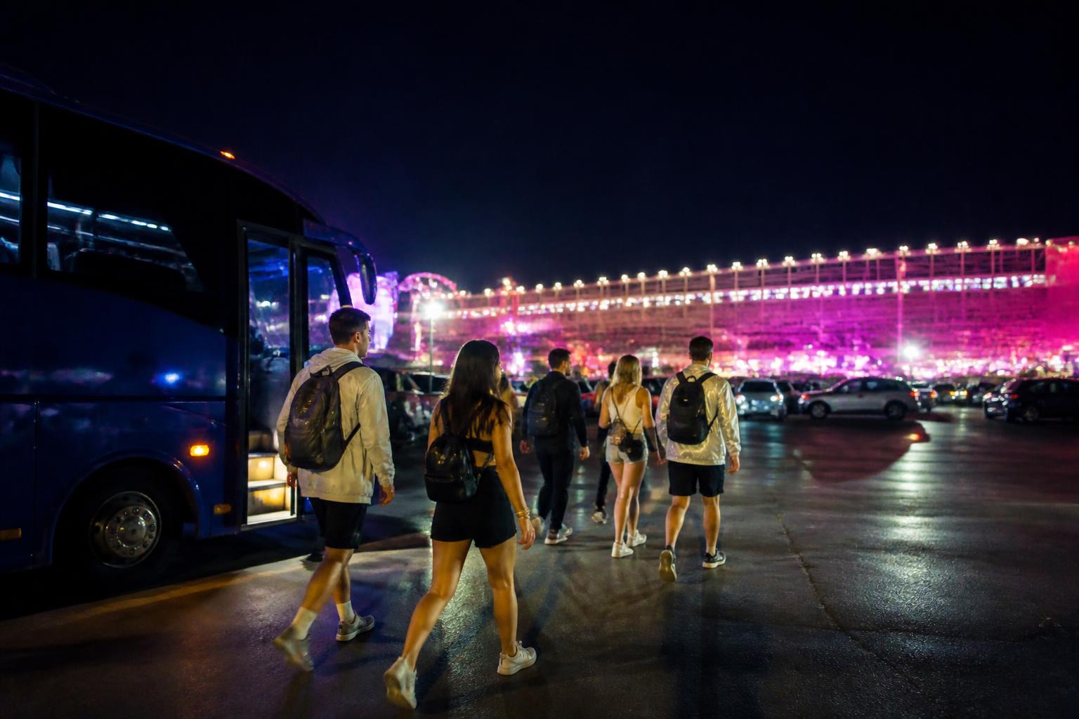 Shuttle bus llegando al Las Vegas Motor Speedway en la noche con travelers caminando hacia la entrada del festival