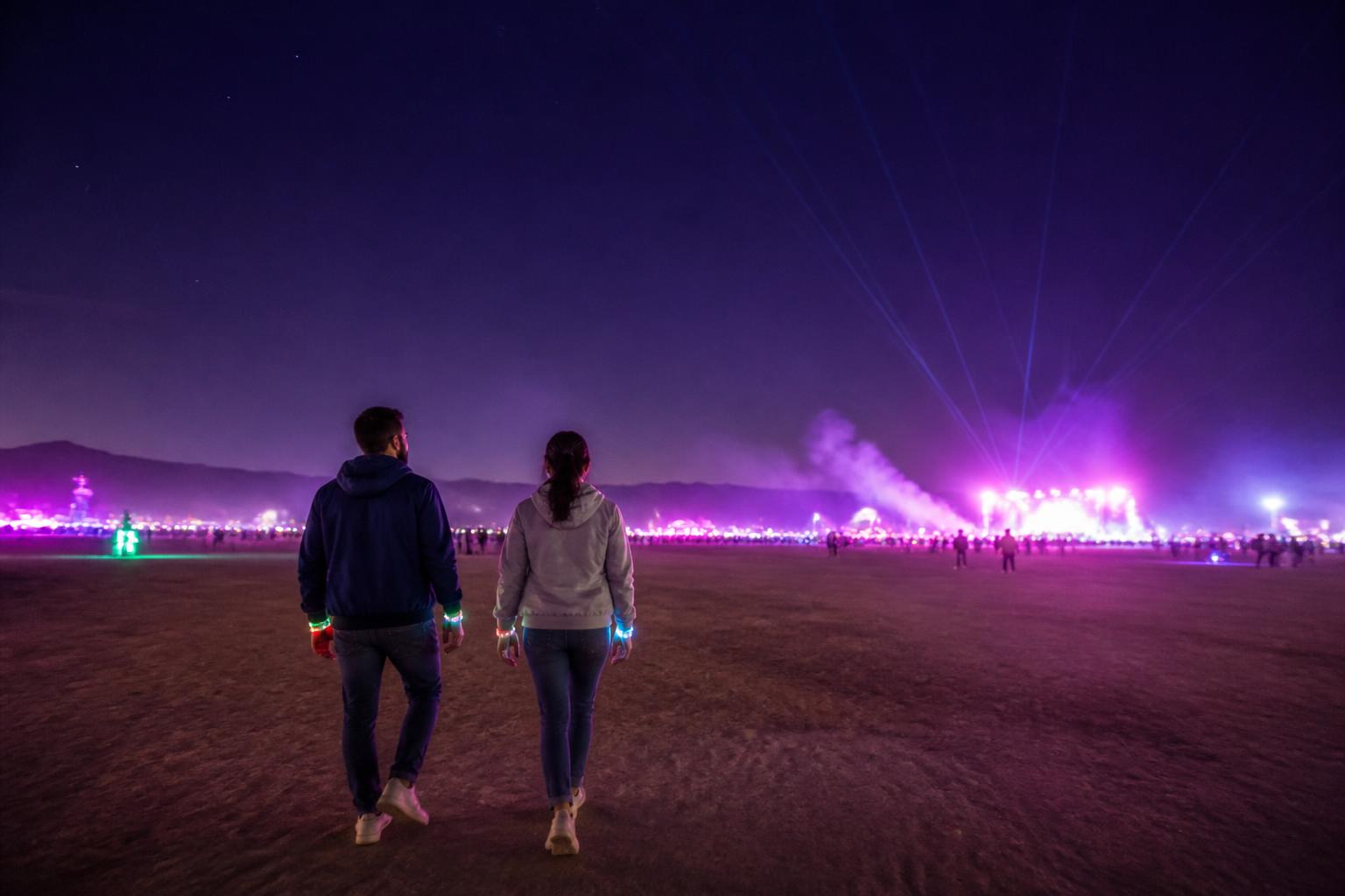 Pareja de travelers con pulseras LED y abrigo liviano caminando bajo el cielo nocturno del desierto de Nevada cerca del speedway