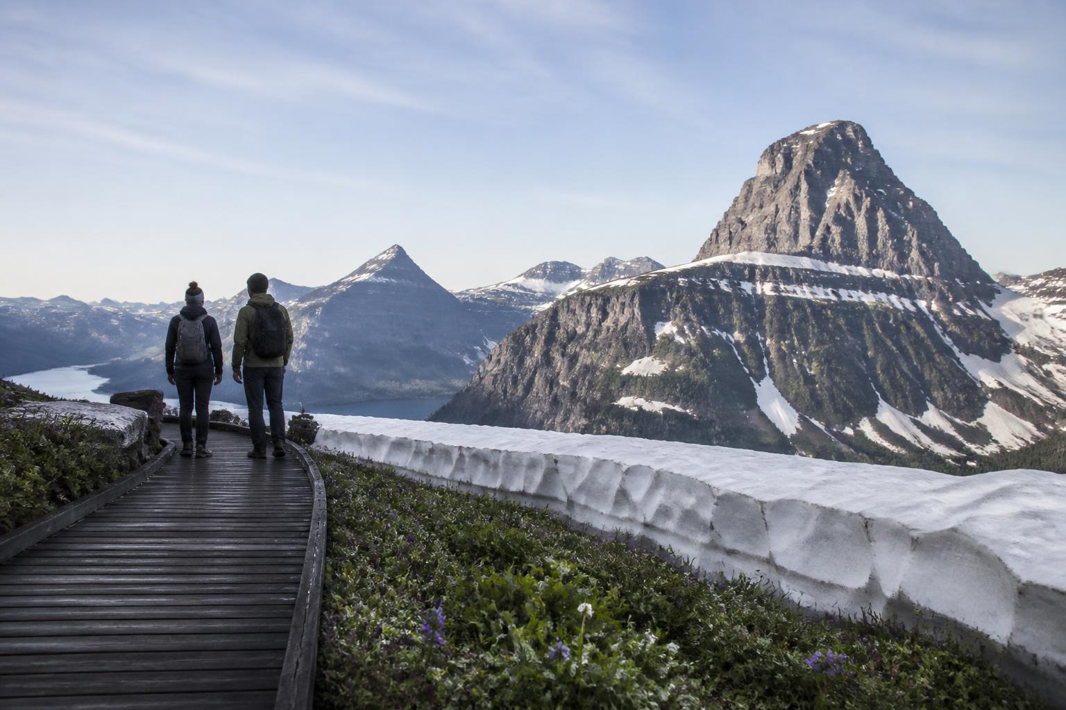 Vista panorámica desde el Logan Pass Visitor Center en Glacier National Park con dos travelers de espalda mirando picos nevados, vegetación alpina con flores silvestres y un cordón blanco al fondo
