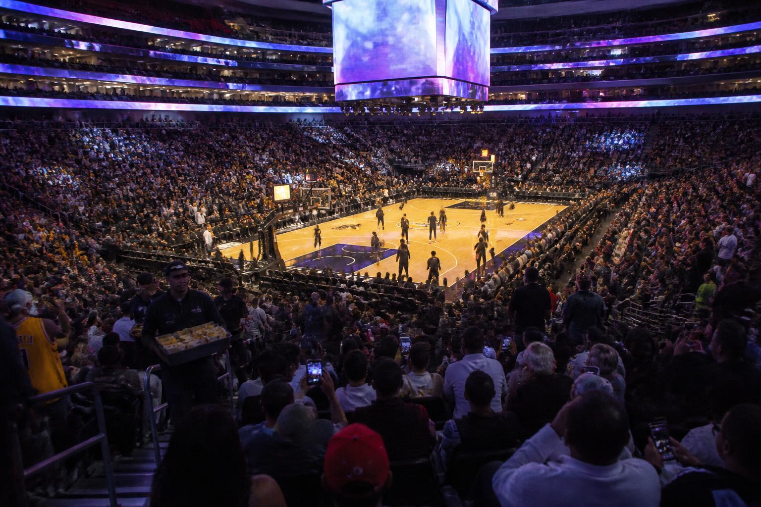 Vista interior de un arena de basquet de la NBA en hora de calentamiento, jugadores en cancha realizando ejercicios previos, pantalla central encendida, gradas iluminadas con miles de hinchas tomando fotos con el celular, atmósfera previa al partido
